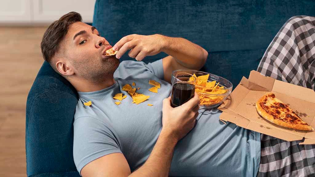 Man eet ongezond op de bank met chips, pizza en frisdrank terwijl hij morst op zijn kleding